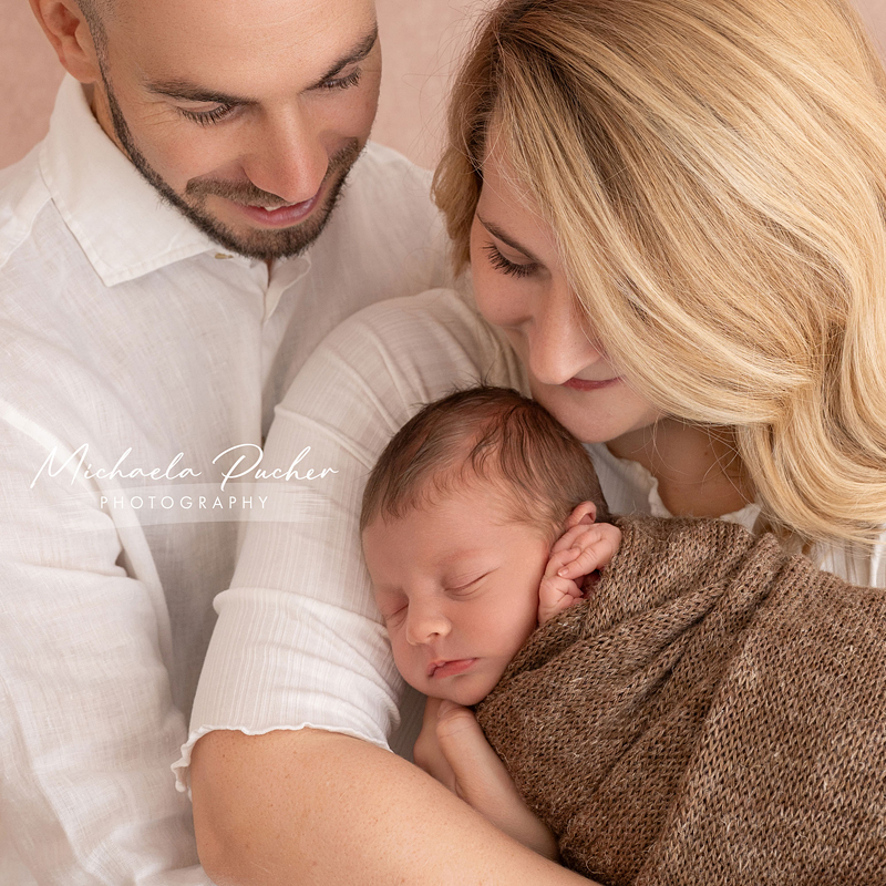 Mama und Papa halten ihr schlafendes Neugeborenes liebevoll im Arm; beide lächeln sanft und schauen beschützend auf das Baby.